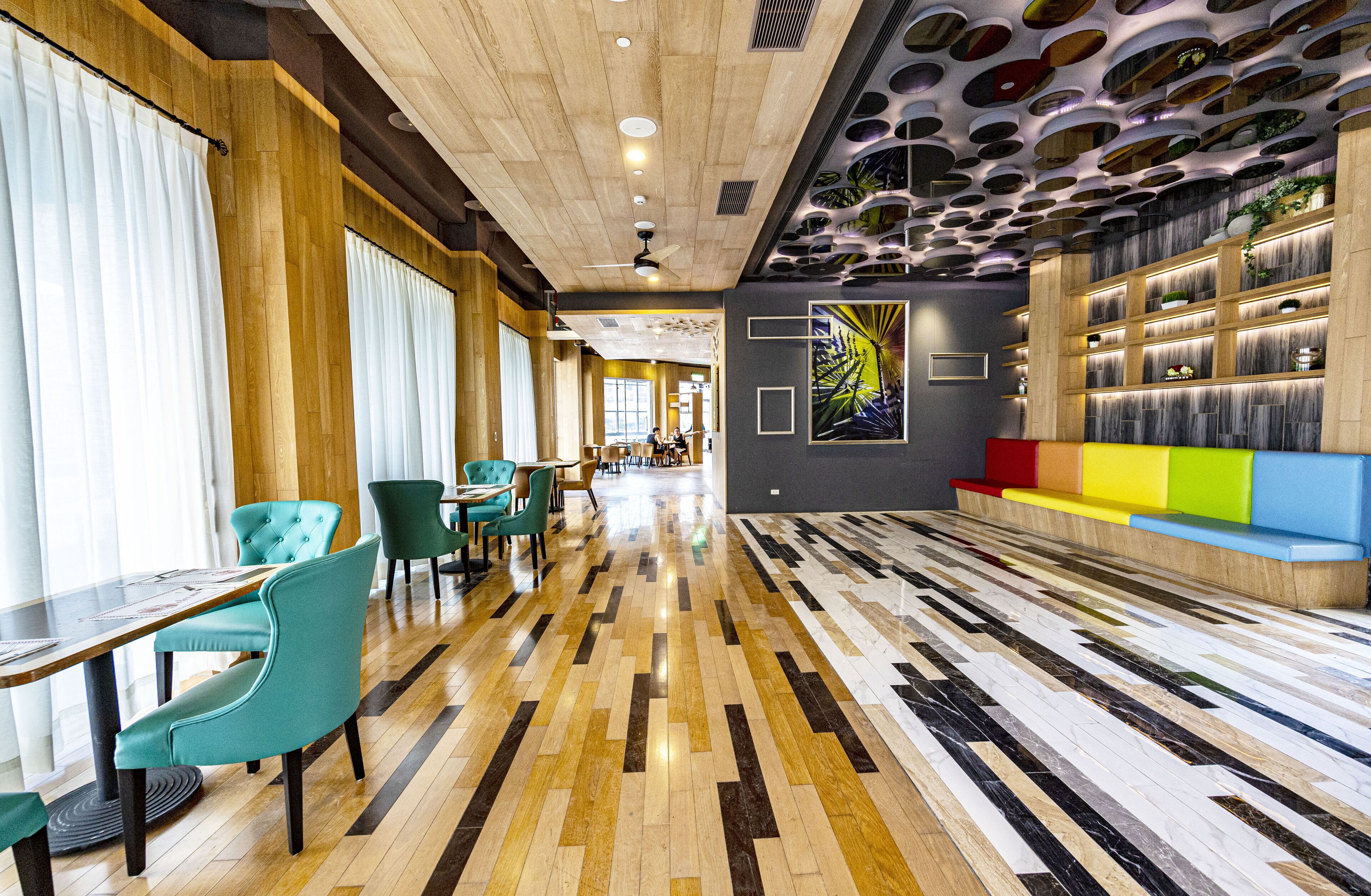 A modern hotel lobby with a wooden ceiling, a patterned floor, teal chairs, and a colorful seating area.