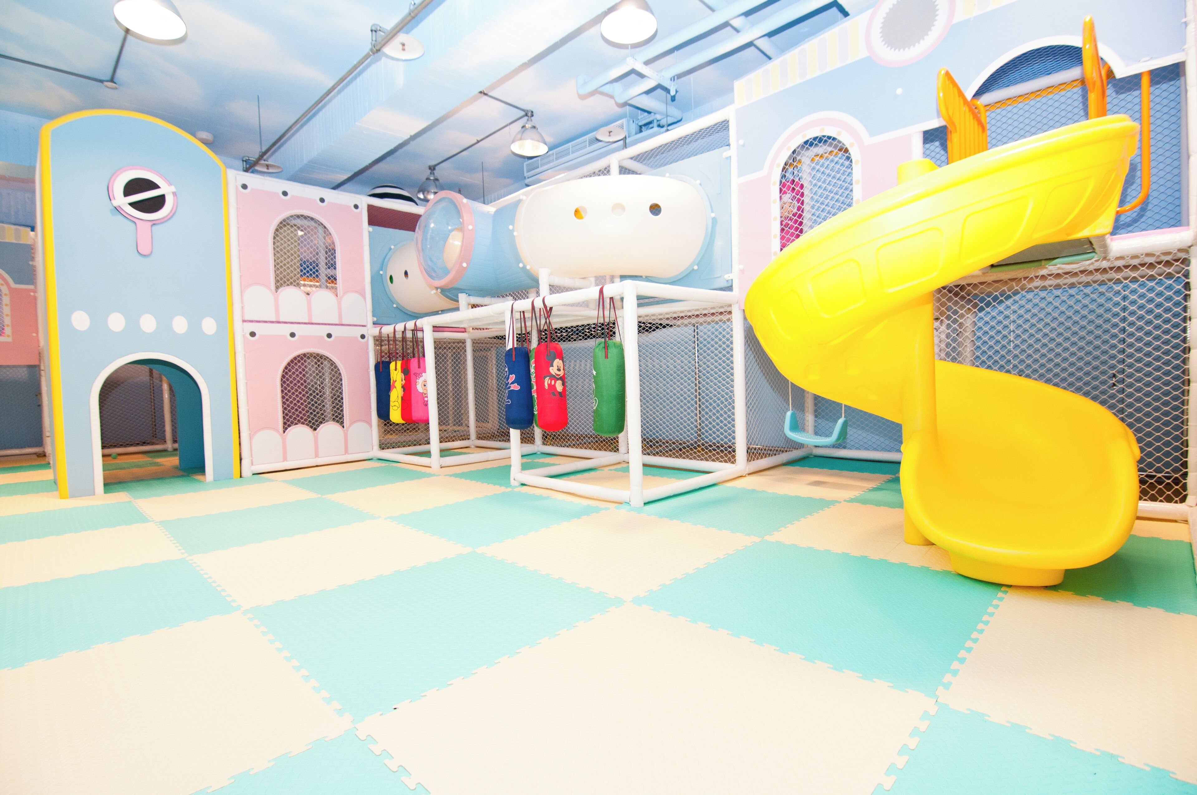 Un espace de jeux pour enfants avec un toboggan jaune, un sol rembourré et des structures de jeu colorées.