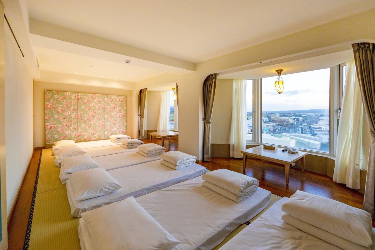 Hotellihuone, jossa on neljä sänkyä, suuri ikkuna, josta on näkymä, pieni pöytä ja koristeellinen seinäpaneeli.