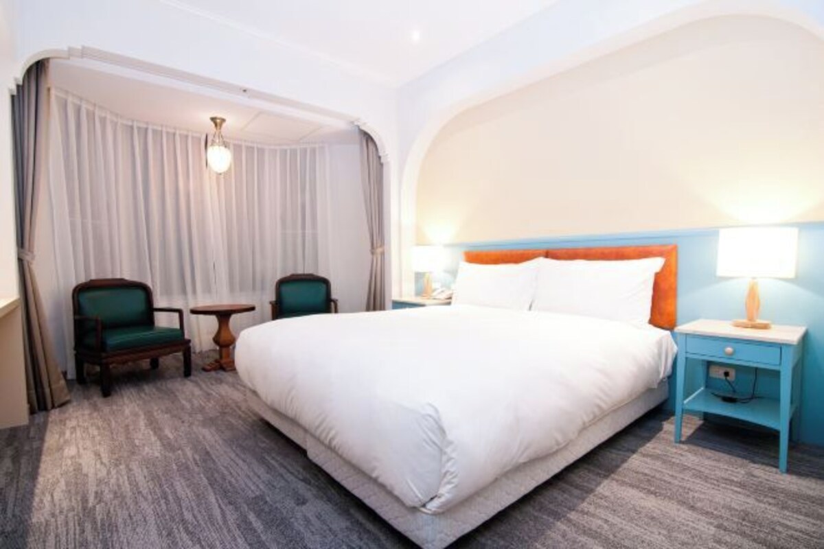 Une chambre d’hôtel avec un grand lit, deux chaises, une petite table et une table de chevet avec une lampe.