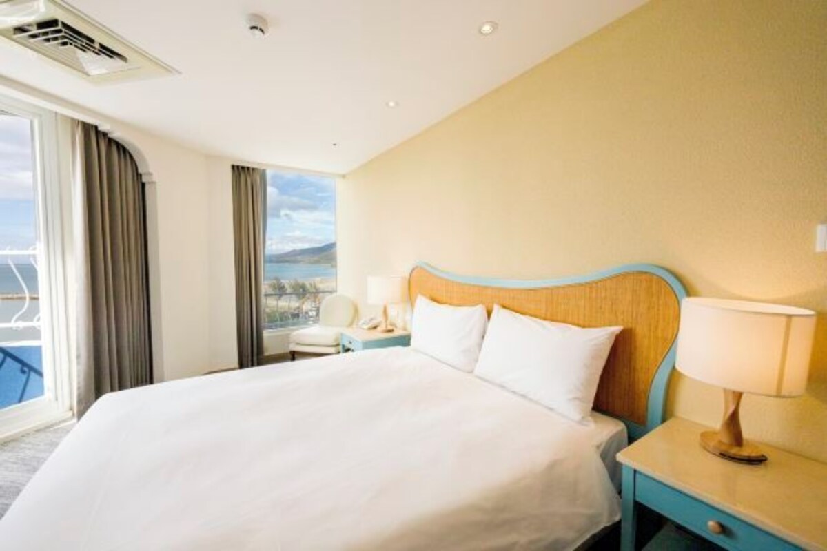 Une chambre d’hôtel avec un grand lit, une table de chevet avec une lampe, une fenêtre avec des rideaux et une vue sur la mer.