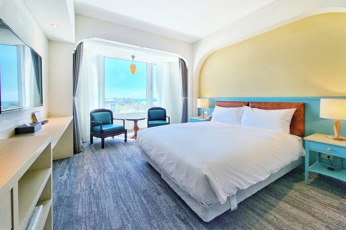 Hotellihuone, jossa on suuri sänky, kaksi tuolia, työpöytä ja televisio.