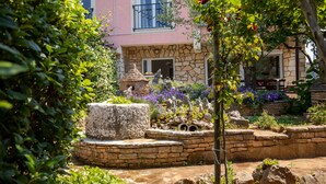 A stone fountain surrounded by lush greenery and a pink building with a balcony. - Flora relax house (Fažana)