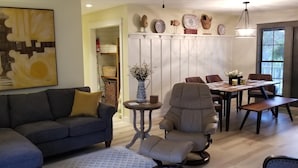 Ein Wohnzimmer mit grauem Sofa, einem runden Tisch mit einer Vase und einem Essbereich mit Holztisch und Stühlen.