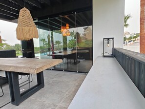 Un espace extérieur aménagé avec une table et des chaises en bois, un luminaire suspendu et un mur de verre donnant sur une piscine et une végétation luxuriante.