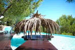 Un área de piscina con cubierta de madera, una estructura de sombrilla de paja y una cascada al fondo.