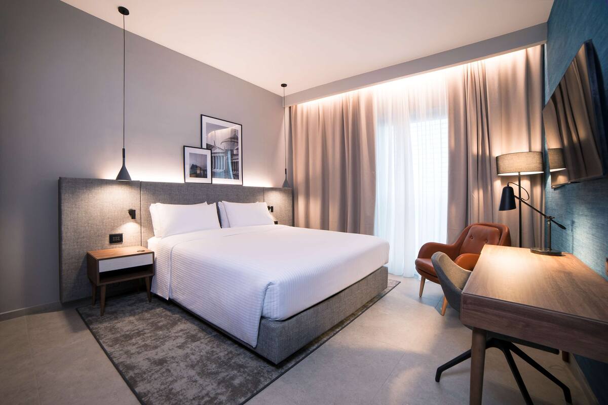 A modern hotel room with a large bed, a desk, a chair, and a nightstand.