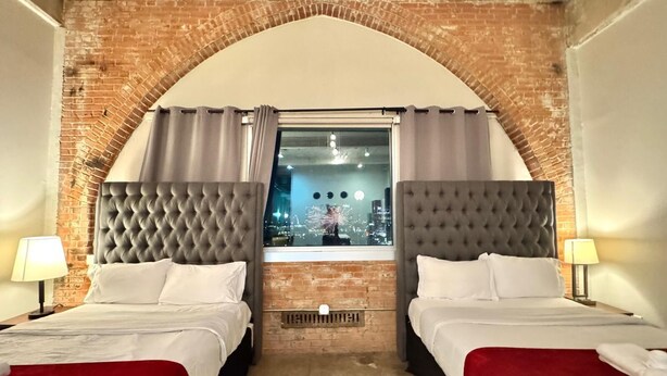 A hotel room with two beds, a brick wall, and a window with a city view.