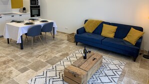 A modern living room with a blue sofa, a dining table set for two, and a kitchen area with white cabinets and a yellow accent wall.