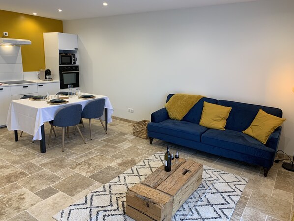 A modern living room with a blue sofa, a dining table set for two, and a kitchen area with white cabinets and a yellow accent wall.