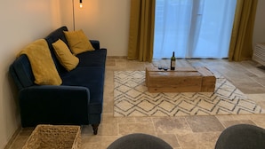 A living room with a dark blue sofa, yellow cushions, a wooden coffee table with a bottle and two glasses, and a patterned rug.