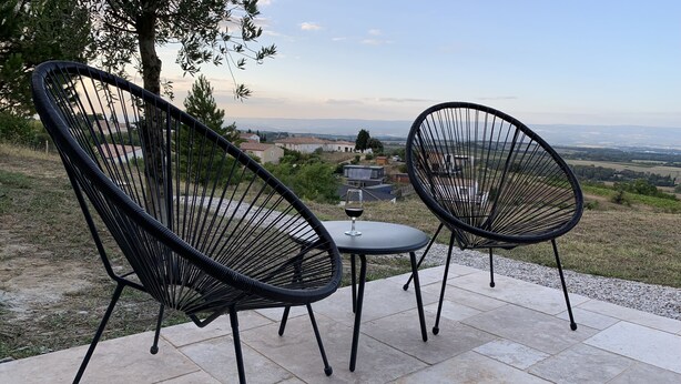 Two black wireframe chairs and a small table with a wine glass on a patio with a view of a distant landscape.