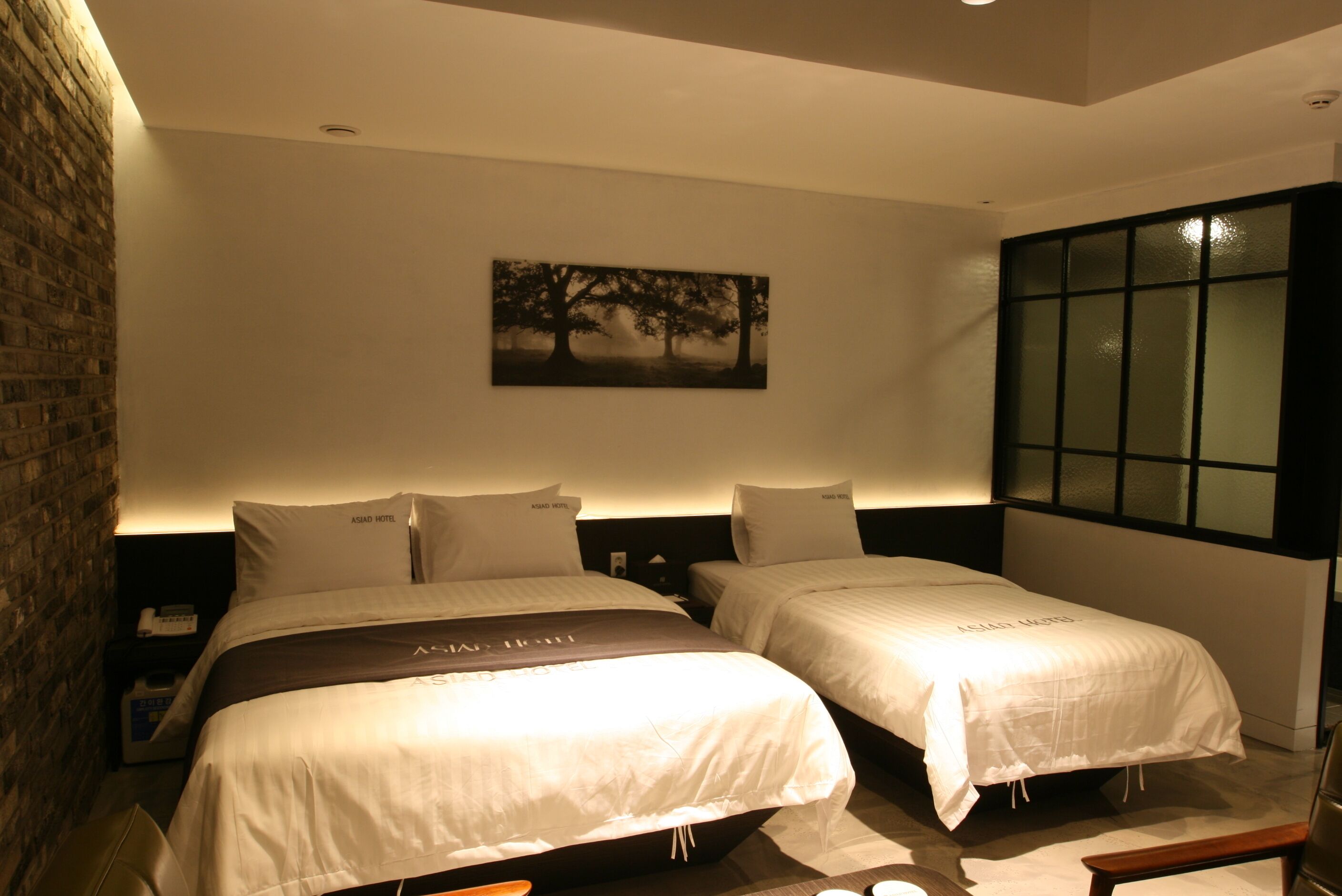 Habitación de hotel con dos camas, un muro de ladrillo, un cuadro y un ventanal grande.