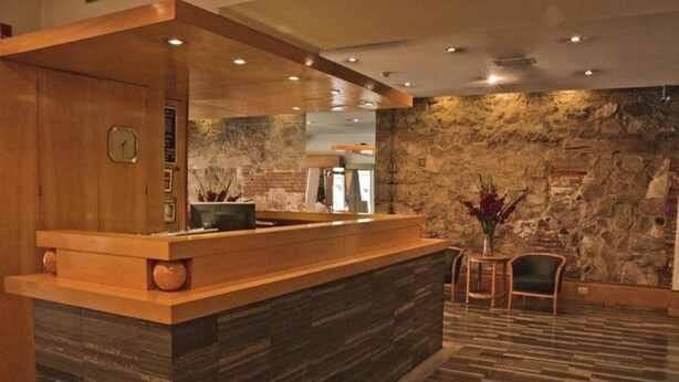 A reception area with a wooden counter, stone wall, and a small seating area with chairs and a table. - Hotel Felipe II (Córdoba)