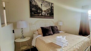 A neatly made bed with a patterned bedspread, two bedside lamps, and a cityscape painting above it. - Cabin on the clouds. (Cocagne)