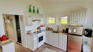 A compact kitchen with white cabinets, a stove, a sink, and a refrigerator. There are three windows providing natural light, and a small countertop with a coffee maker and a kettle. - Cabin on the clouds. (Cocagne)