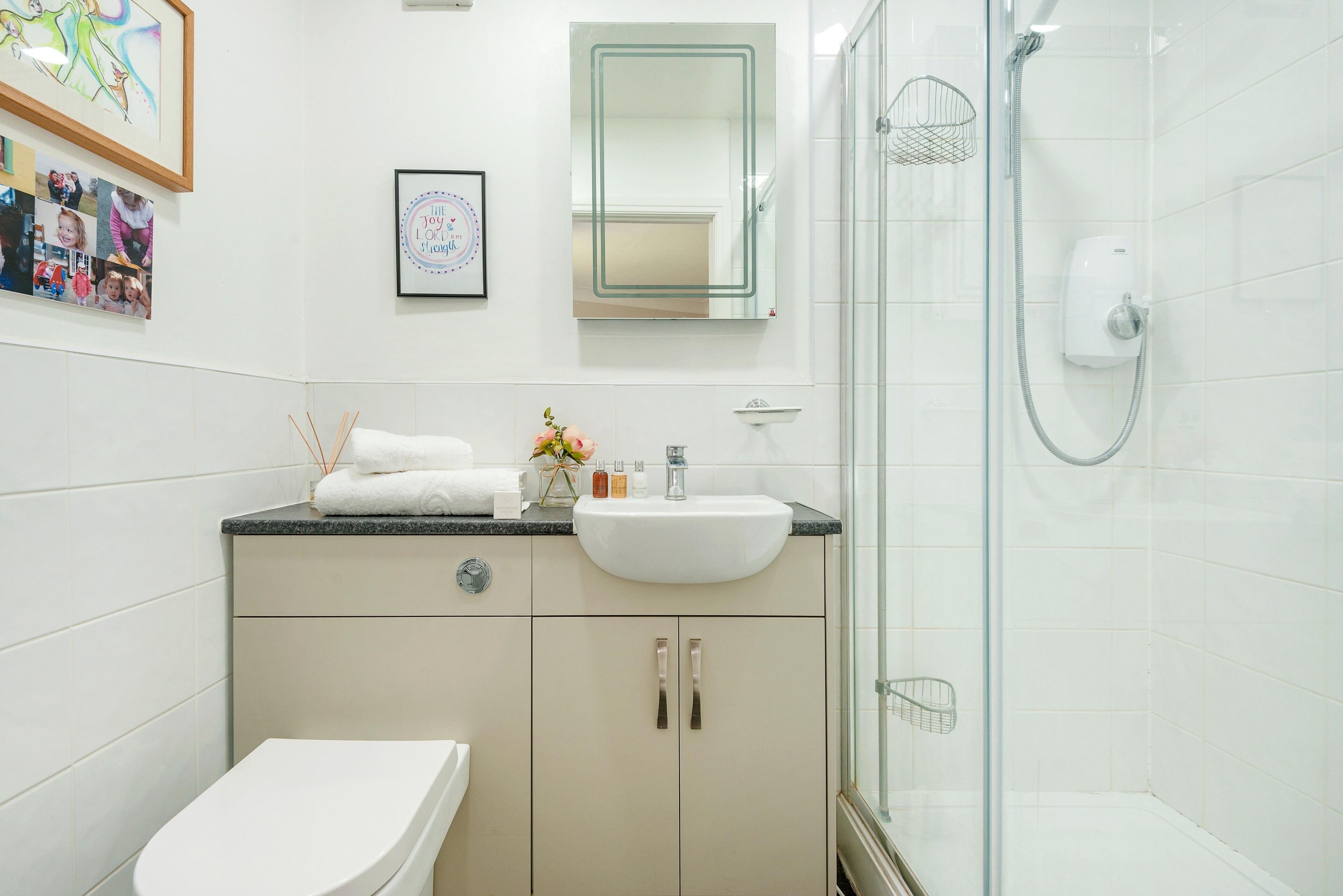 A modern bathroom with a white sink, beige cabinets, a glass shower enclosure, and a framed photo collage on the wall.