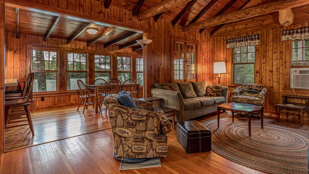 A wooden cabin interior with a large dining table, two sofas, and a fireplace.