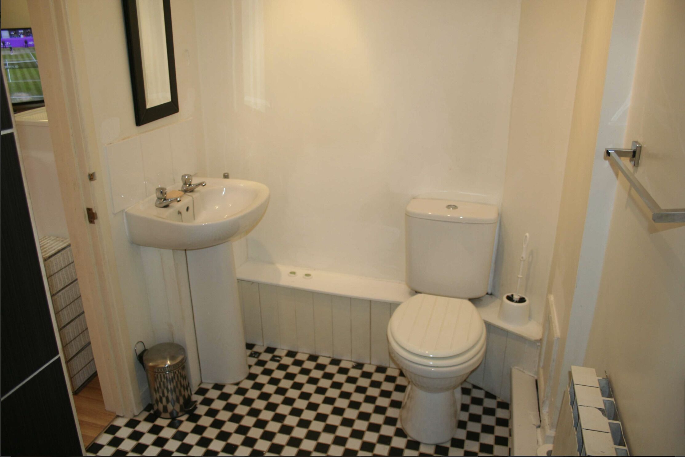 Une salle de bain avec un lavabo, des toilettes et une baignoire, tous de couleur blanche. Le sol est recouvert d’un motif à carreaux noirs et blancs.