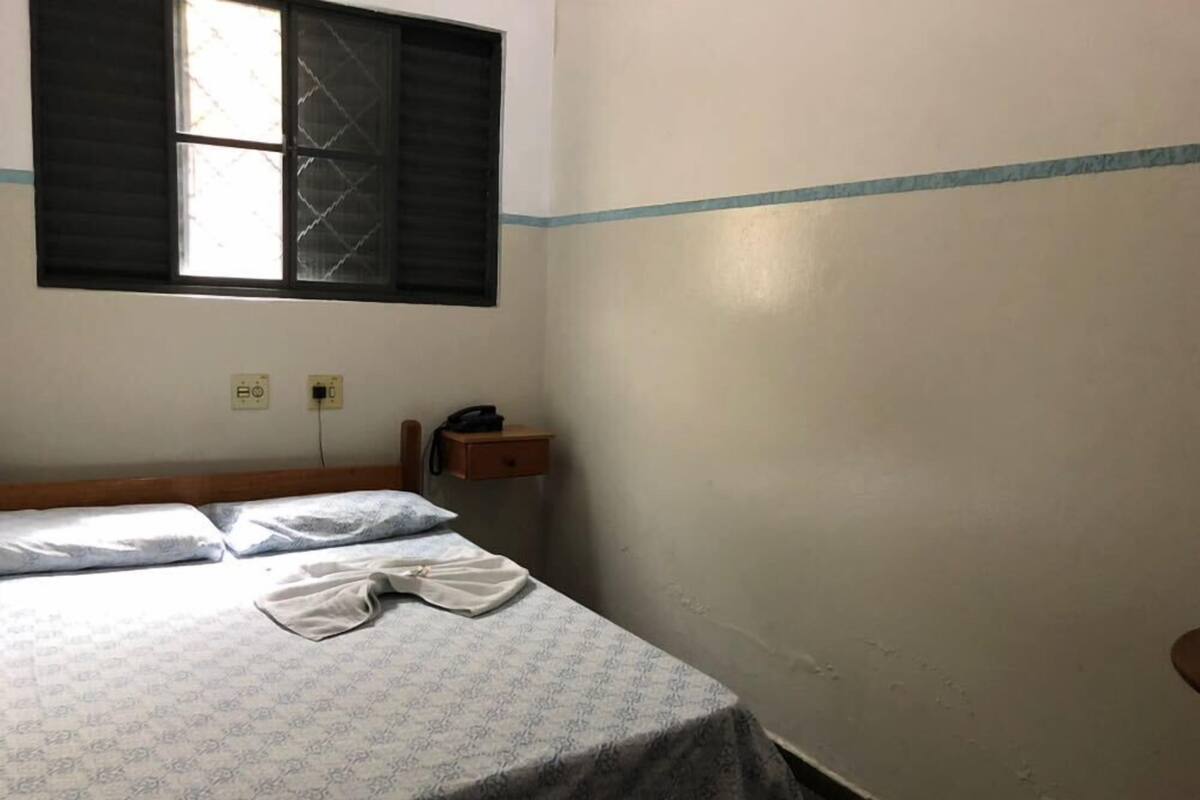 Un lit simple avec une couvre-lit à motifs, un téléphone sur la table de chevet et une fenêtre dont les stores sont fermés.
