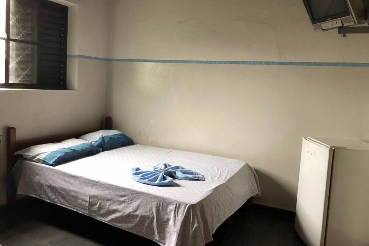 Un lit simple avec un oreiller à rayures bleues et blanches et une serviette pliée, une fenêtre avec des stores et un petit téléviseur fixé au mur.