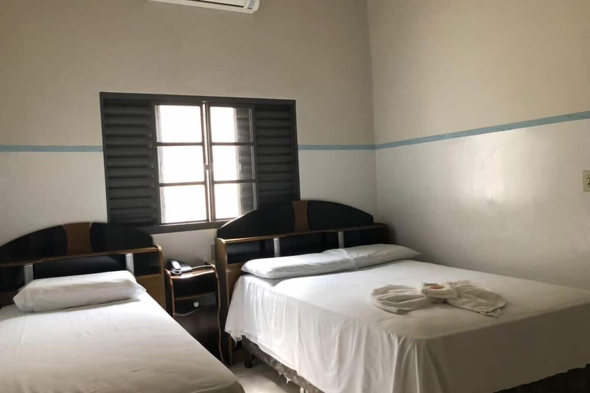 Une chambre avec deux lits simples, une petite table de chevet avec une lampe, une fenêtre avec des volets et un climatiseur fixé au mur.