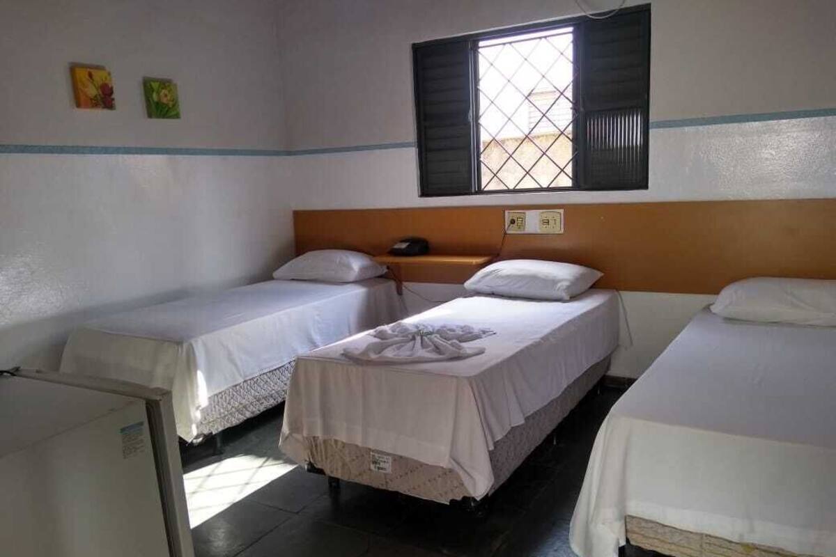 Une chambre avec deux lits simples, une petite table, une fenêtre avec volets et une prise électrique fixée au mur.