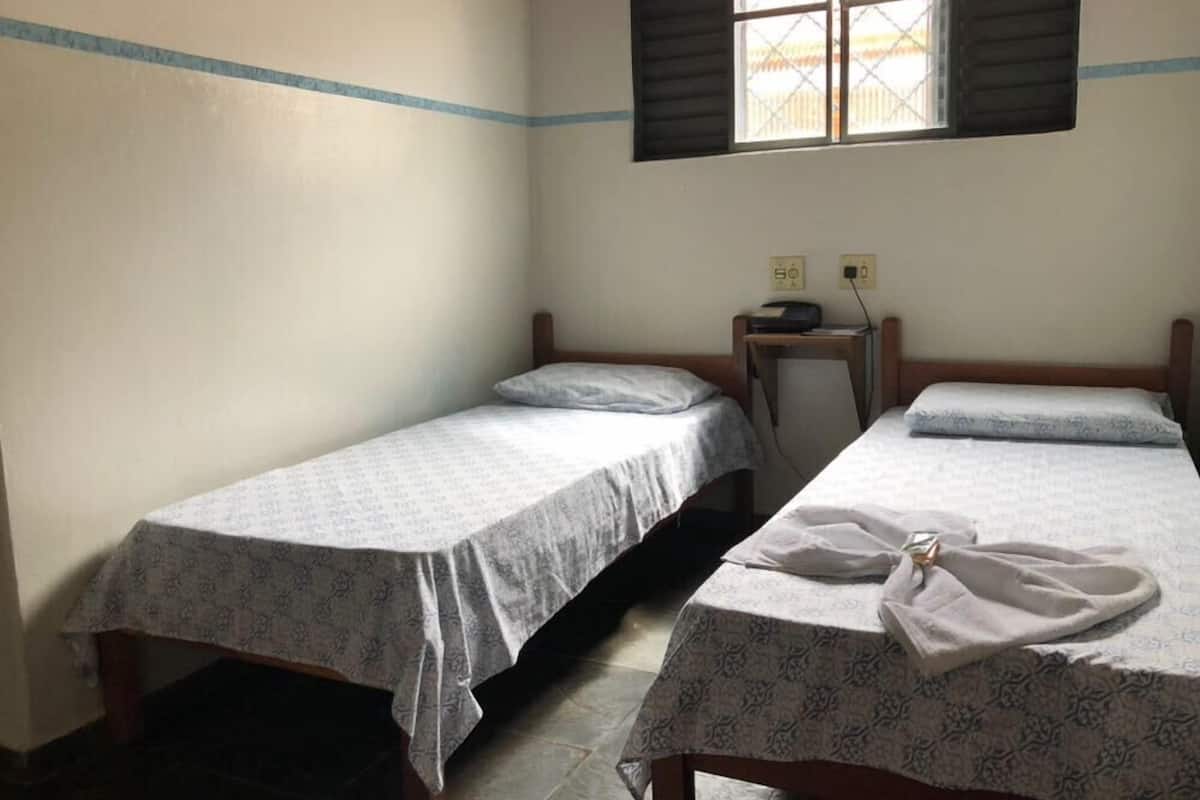 Une chambre avec deux lits simples, une petite table avec une radio et une fenêtre avec des volets.