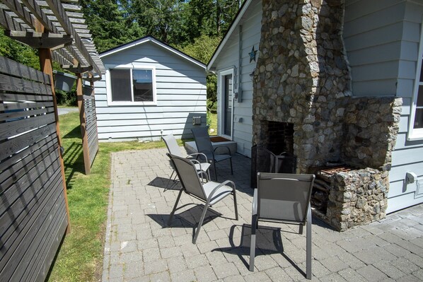 A patio with two chairs and a table, a stone fireplace, and a small house in the background. - Orcas Island, Il Cheto #279 (Orcas Island)