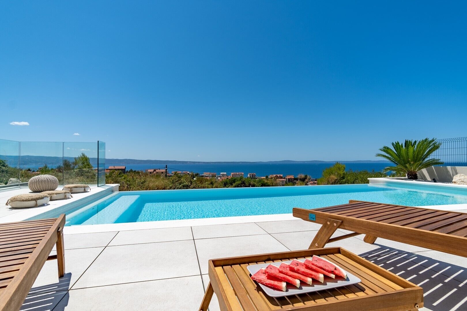 Un espace aménagé au bord de la piscine, offrant une vue dégagée sur la mer, avec des chaises longues en bois et un plateau de tranches de pastèque.