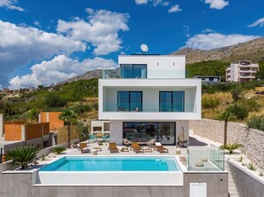 A modern two-story house with a swimming pool, outdoor seating, and a view of the surrounding landscape.