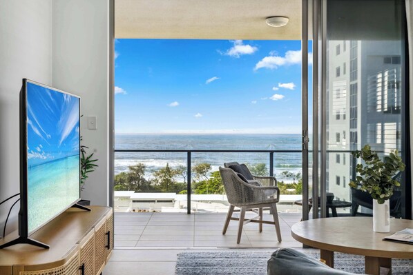 Un salon moderne avec un grand téléviseur, un balcon offrant une vue sur l’océan, un fauteuil en osier et une table ronde.