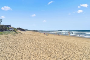 A wide sandy beach with a few people and beach umbrellas, a lifeguard tower, and a building on the left. - Cotton Tree Ocean Views +  Romantic Getaway (Maroochydore)