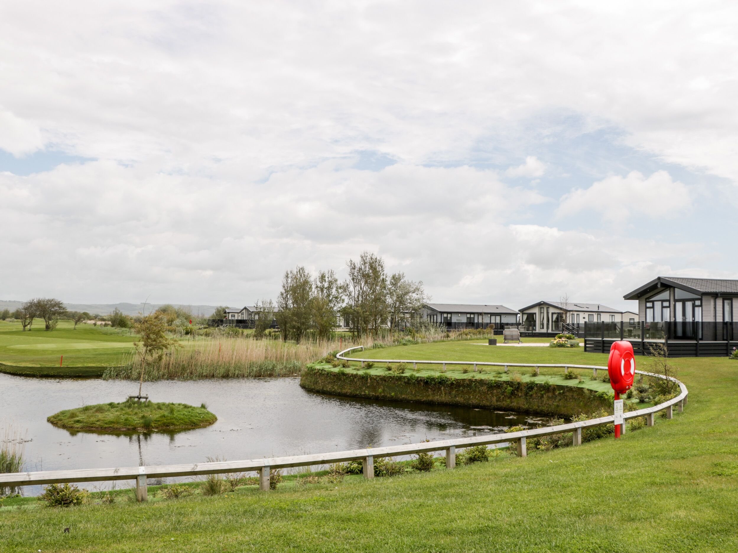 A golf course with a pond, a wooden fence, and modern buildings in the background.