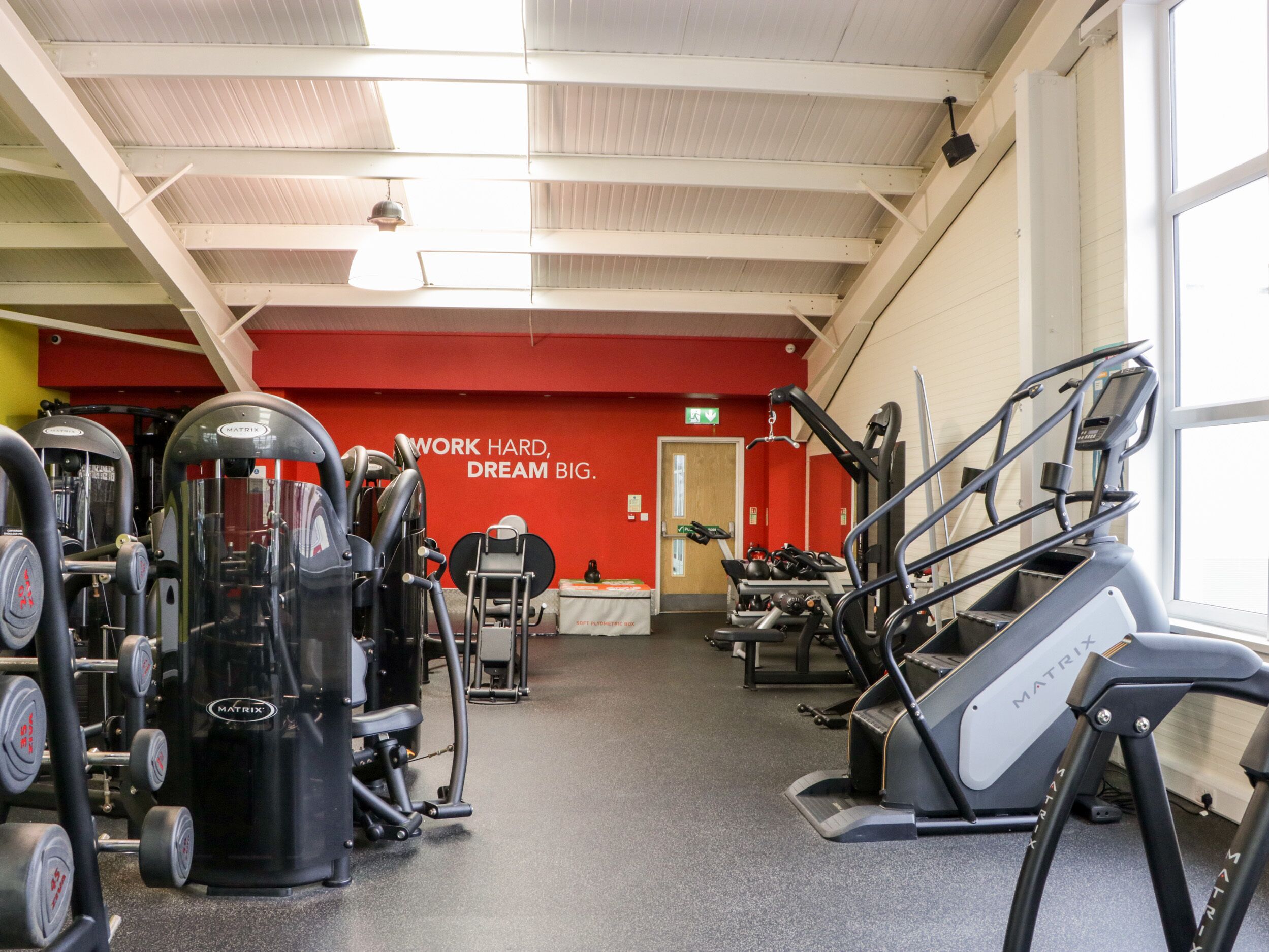 Academia com diversos equipamentos de exercícios, uma parede vermelha com mensagens motivacionais e uma janela que proporciona luz natural.