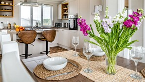 Une cuisine moderne avec des meubles blancs, un coin repas avec une table dressée pour deux personnes et un vase rempli de fleurs.