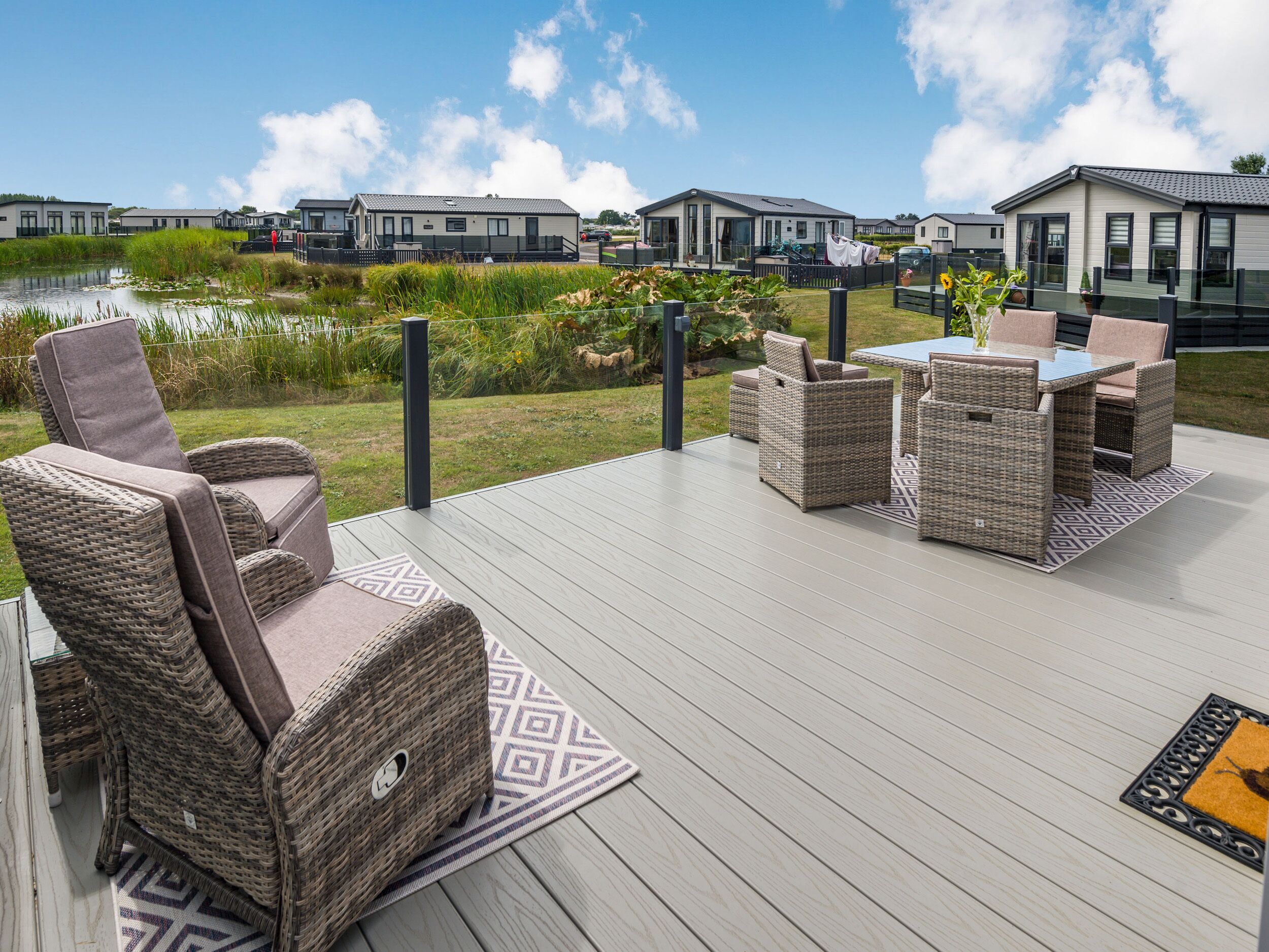 A deck with wicker furniture, a table, and chairs, overlooking a water body and modern buildings.