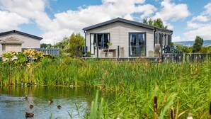 Une maison moderne dotée d’une grande fenêtre et d’une terrasse donnant sur un étang où nagent des canards.