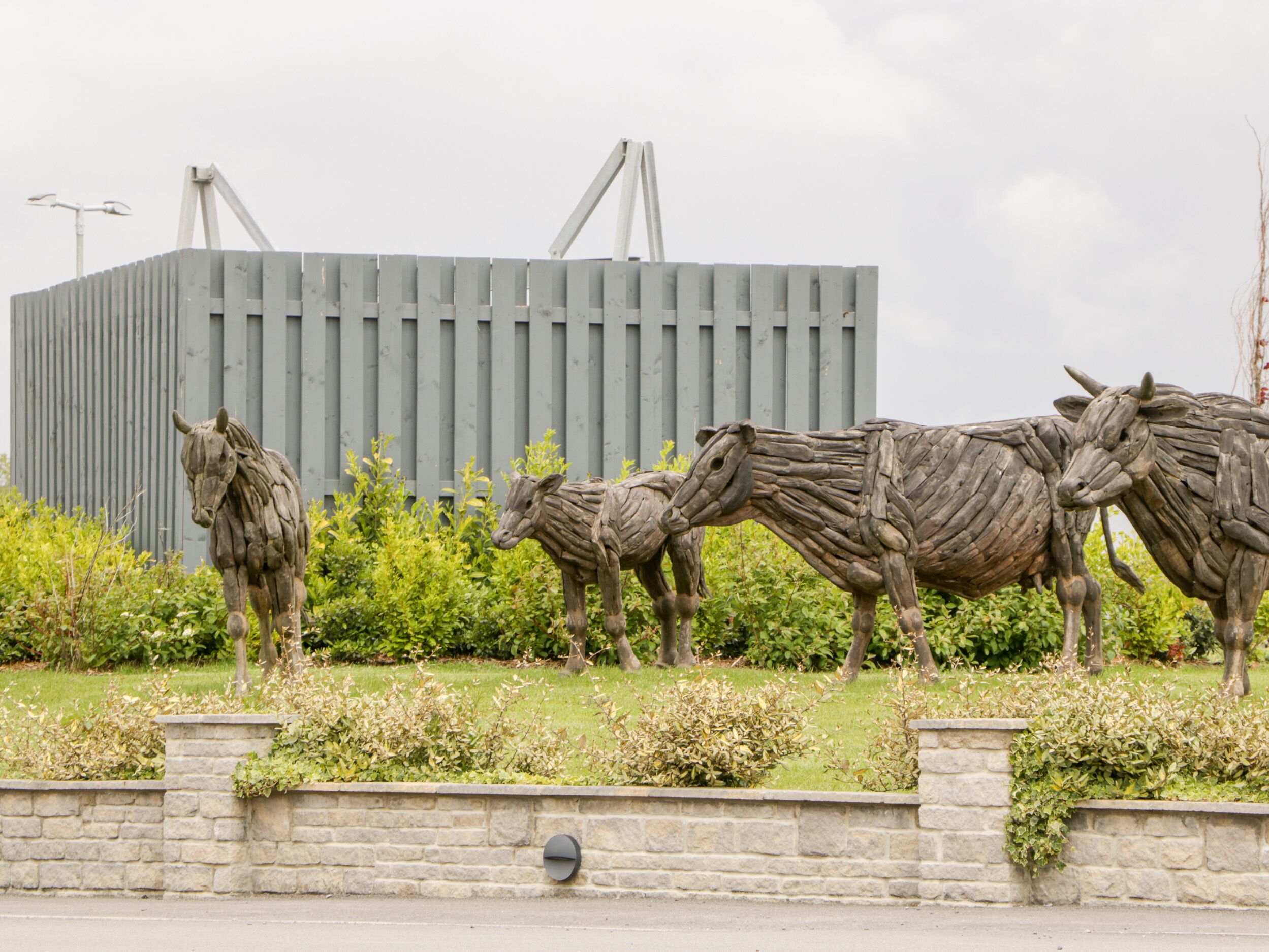 A sculpture of three horses made of wood, with a stone wall and greenery in the foreground, and a modern building in the background.