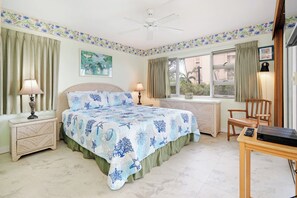 A bedroom with a bed, two bedside tables, a desk, a chair, a ceiling fan, and a window with curtains. - Menehune 127 Partial Ocean Views in Well Maintained Condo Home (Kihei)