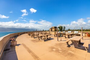 An outdoor patio with picnic tables and barbecue grills, overlooking a beach. - Menehune 127 Partial Ocean Views in Well Maintained Condo Home (Kihei)
