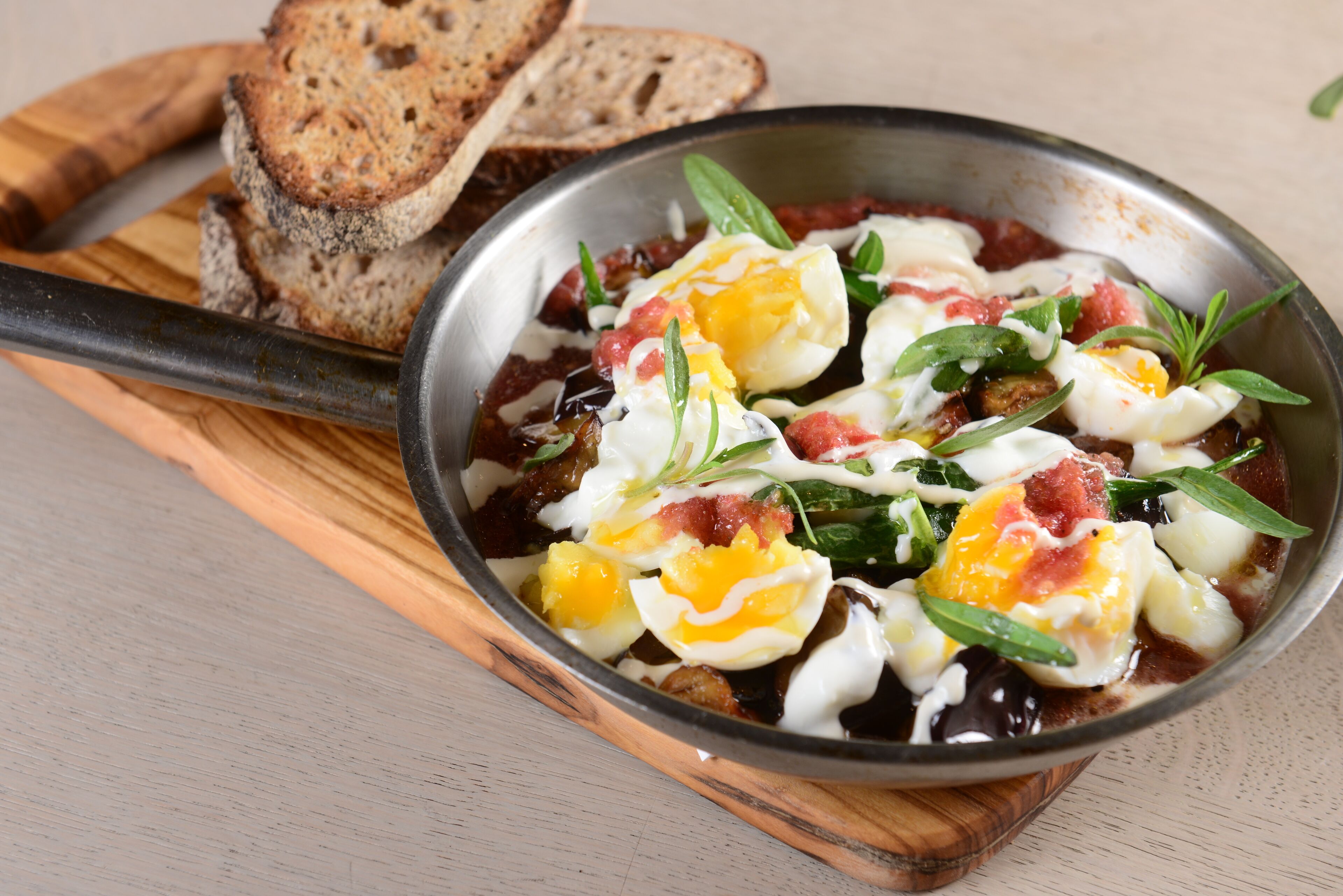 A skillet with eggs, tomatoes, and greens, accompanied by slices of bread on a wooden board.