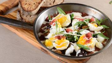 A skillet with eggs, tomatoes, and greens, accompanied by slices of bread on a wooden board.