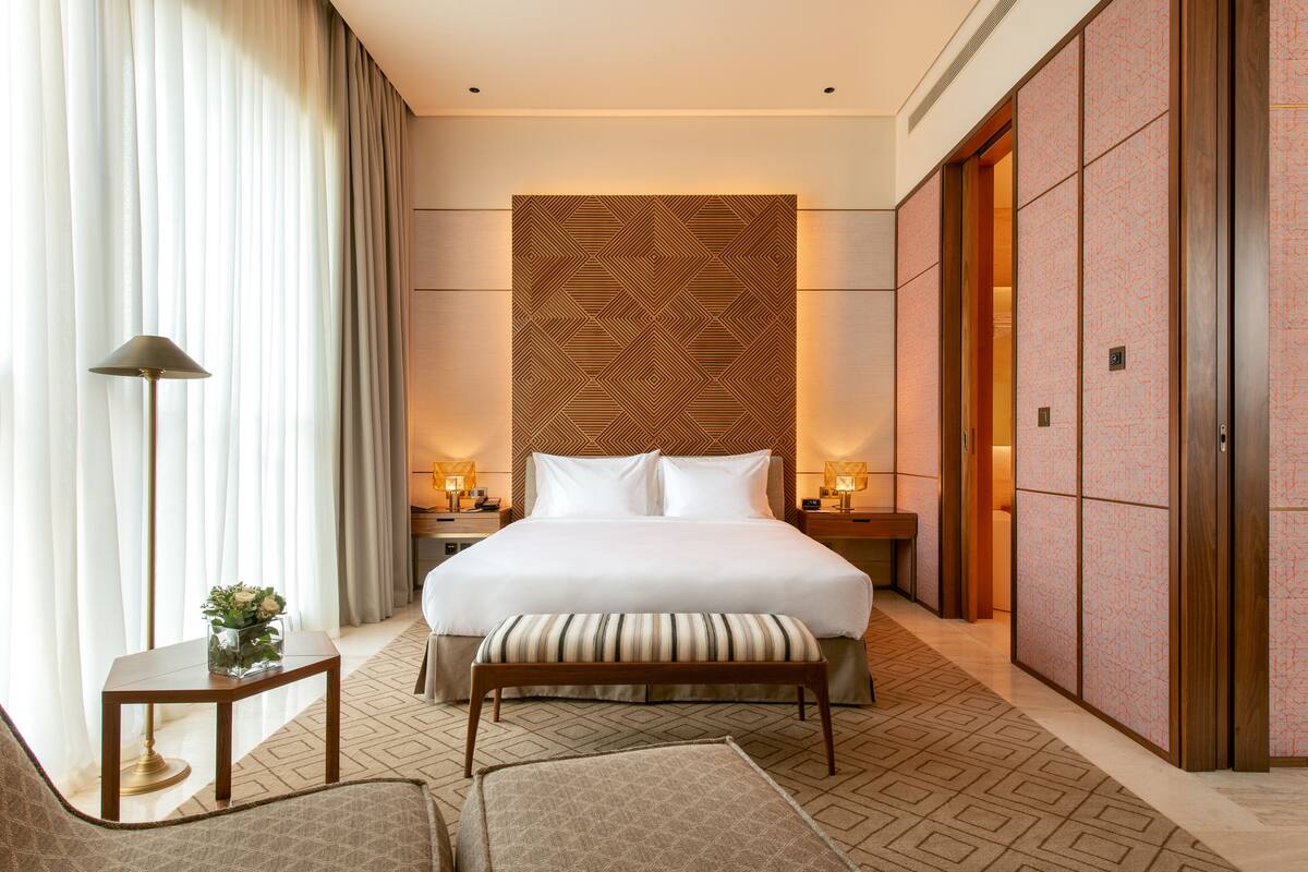 A hotel room with a large bed, a bedside table with a lamp, a bench, and a wardrobe.