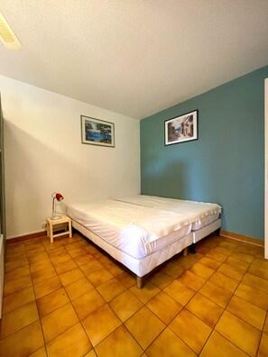 Een kleine eenpersoonskamer met betegelde vloer, nachtkastje met lamp en twee ingelijste foto's aan de muur.