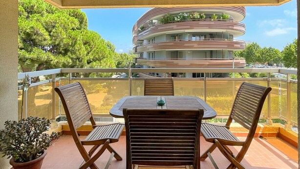 A balcony with a table and two chairs, a potted plant, and a view of a multi-story building with balconies.
