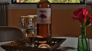 Een fles wijn, twee glazen en een vaas met bloemen op een tafel bij een raam met uitzicht op bomen.