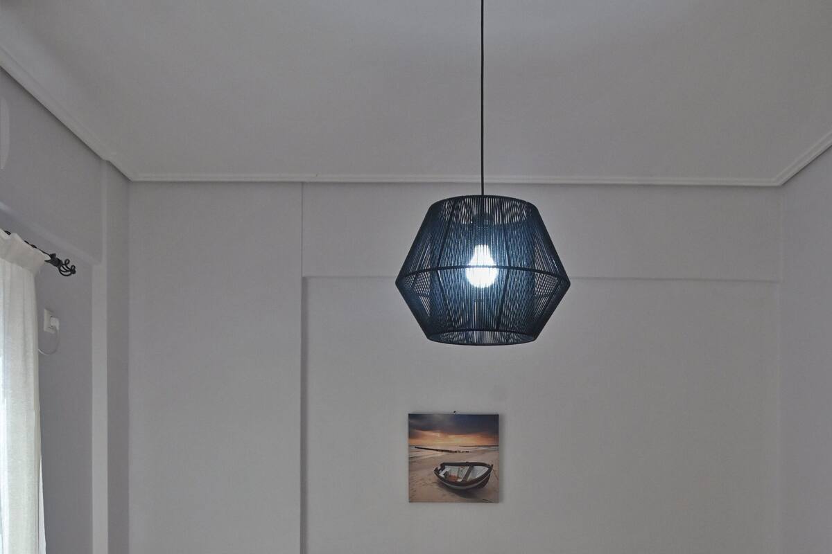 Una cama bien hecha con cabecera gris, una mesita de noche con lámpara y un cuadro en la pared.