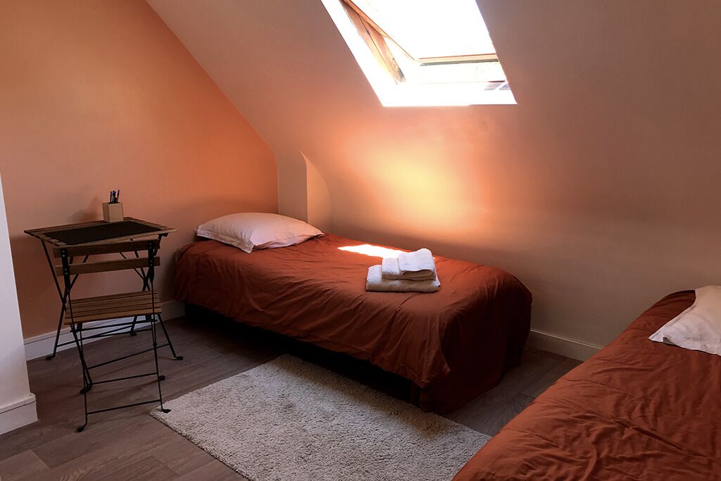 A small, cozy room with a sloped ceiling and a skylight, featuring two beds with matching bedding, a small table with a chair, and a white rug.