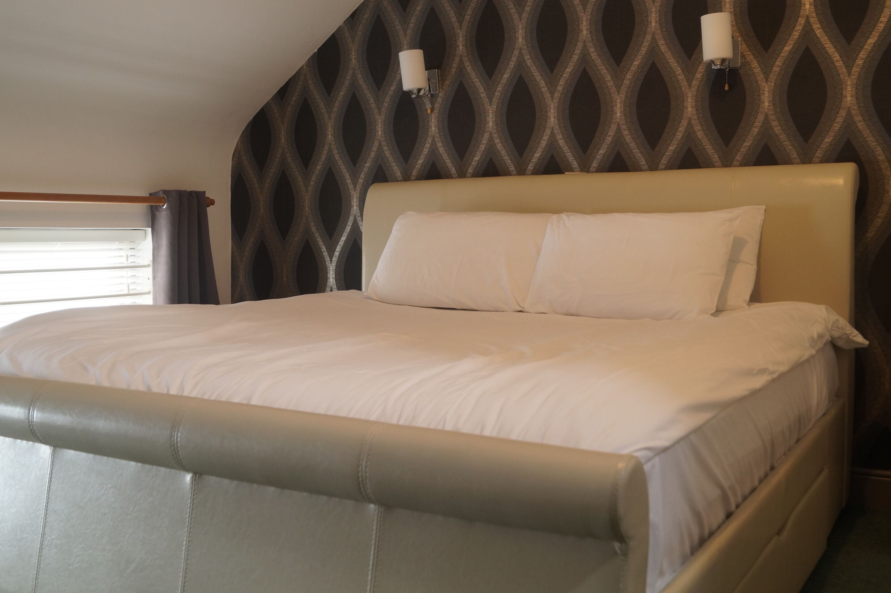 A bed with a headboard, a wall with a geometric pattern, and a window with curtains.
